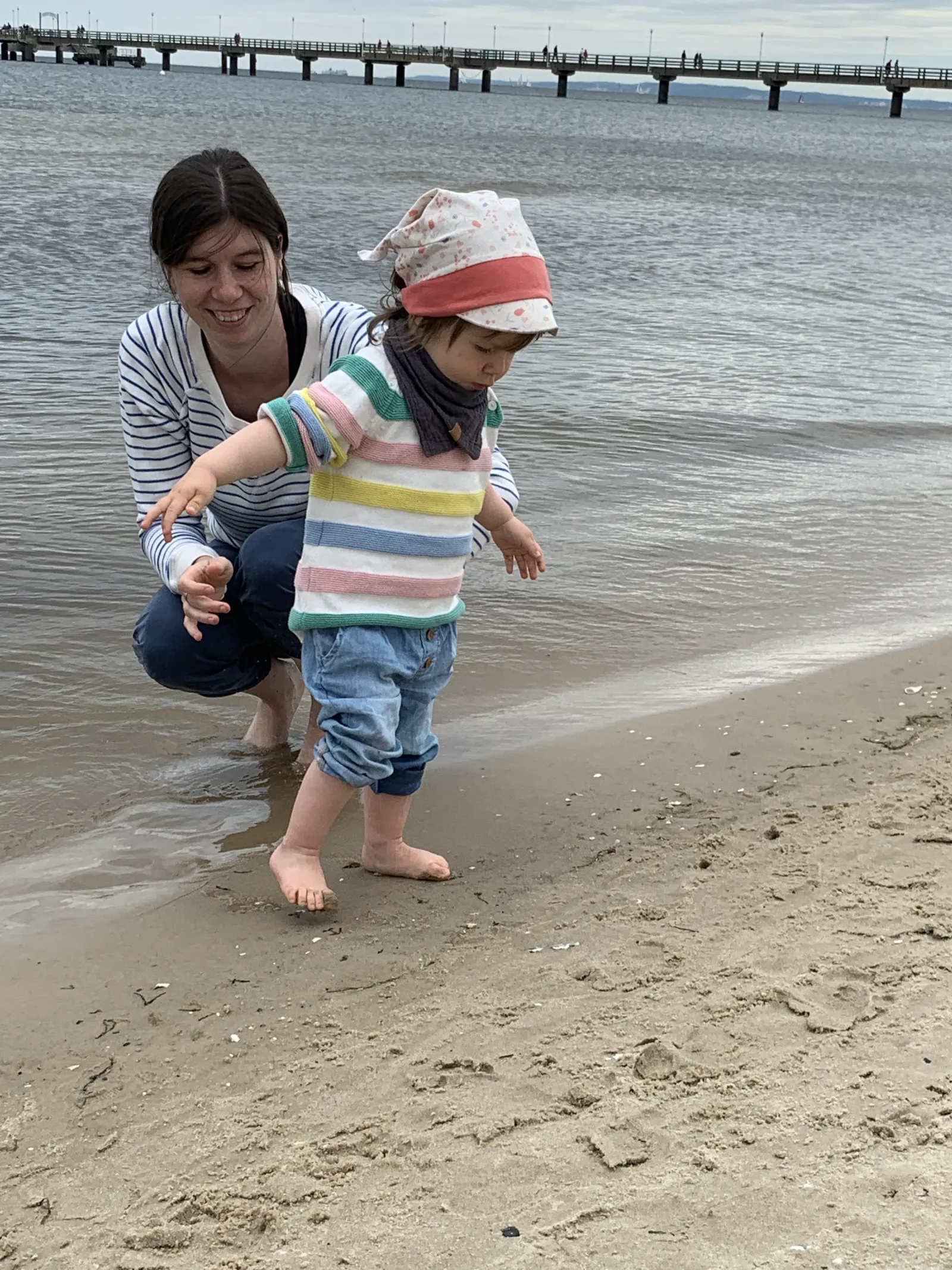 Jule mit Kleinkind am Wassersaum eines Ostseestrands, im Hintergrund die Seebrücke.