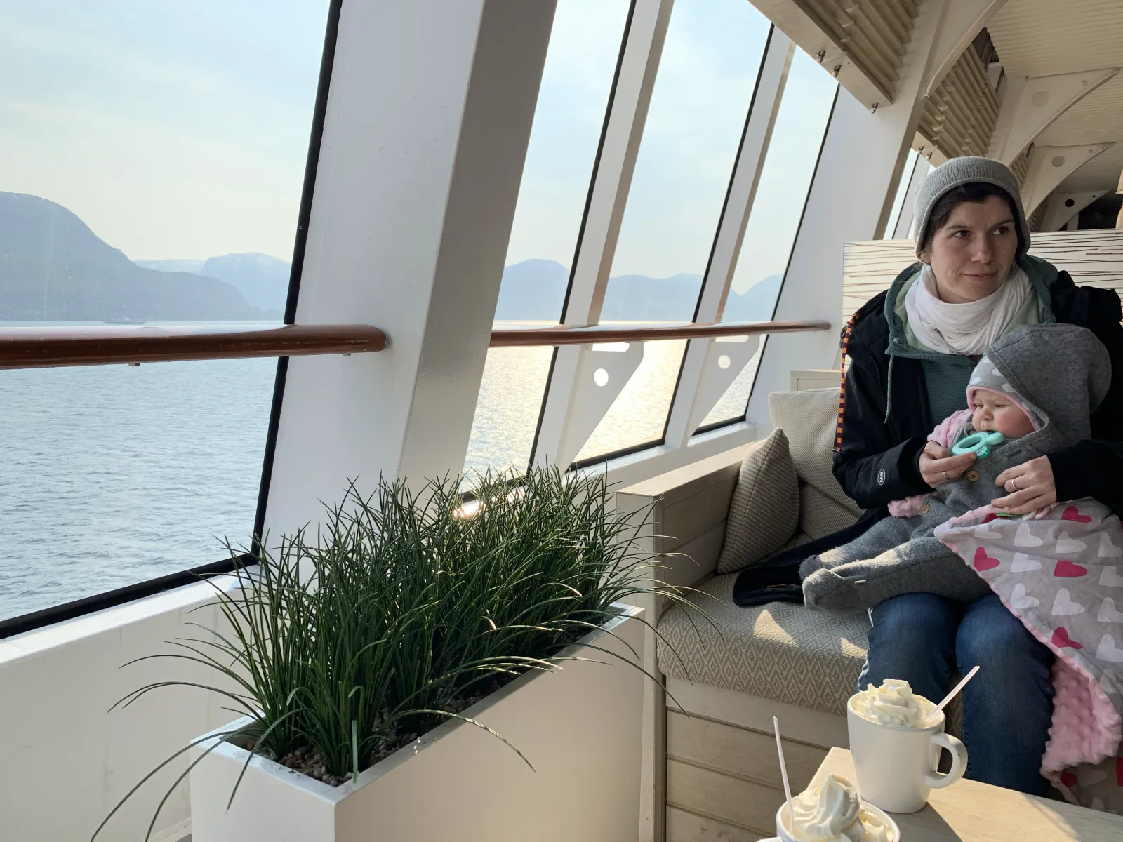 Jule sitzt mit Baby am Panoramafenster eines Schiffs, draußen zieht ein Fjord vorbei.